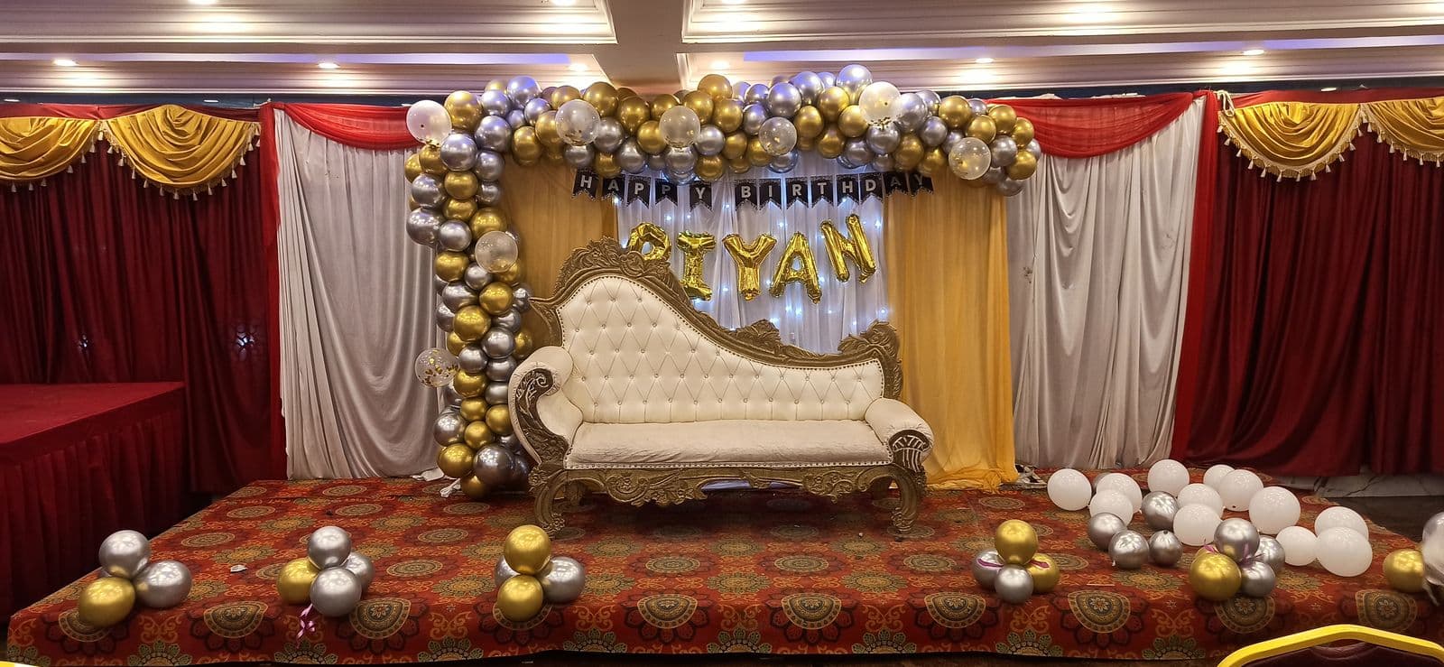 Function hall stage with gold, white and silver balloon arch and white chaise