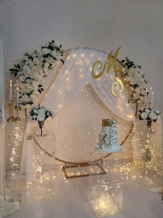 Gold ring backdrop with white roses, fairy-light curtain and monogram behind an engagement cake table